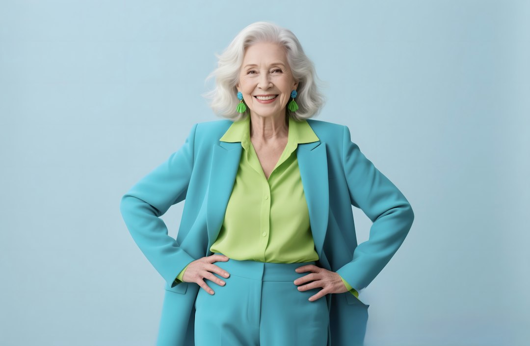 A stylish elderly woman poses in a blue suit and lime green shirt. She exudes confidence with her hands on her hips, against a simple light blue background.
