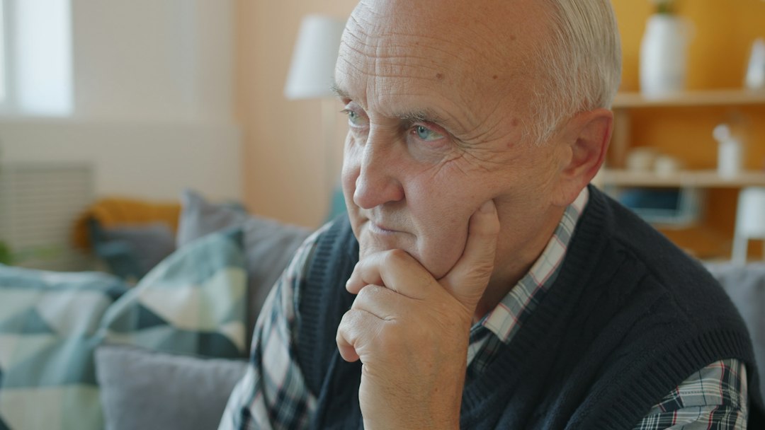 Close-up portrait of serious senior man sitting on sofa at home thinking touching face with hand. Elderly people, retirement and emotions concept.