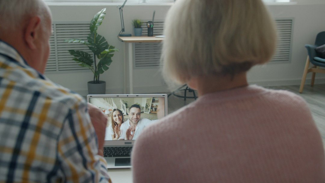 Back view of senior couple making online video call with laptop from home talking to young people using modern device. Family and communication concept.