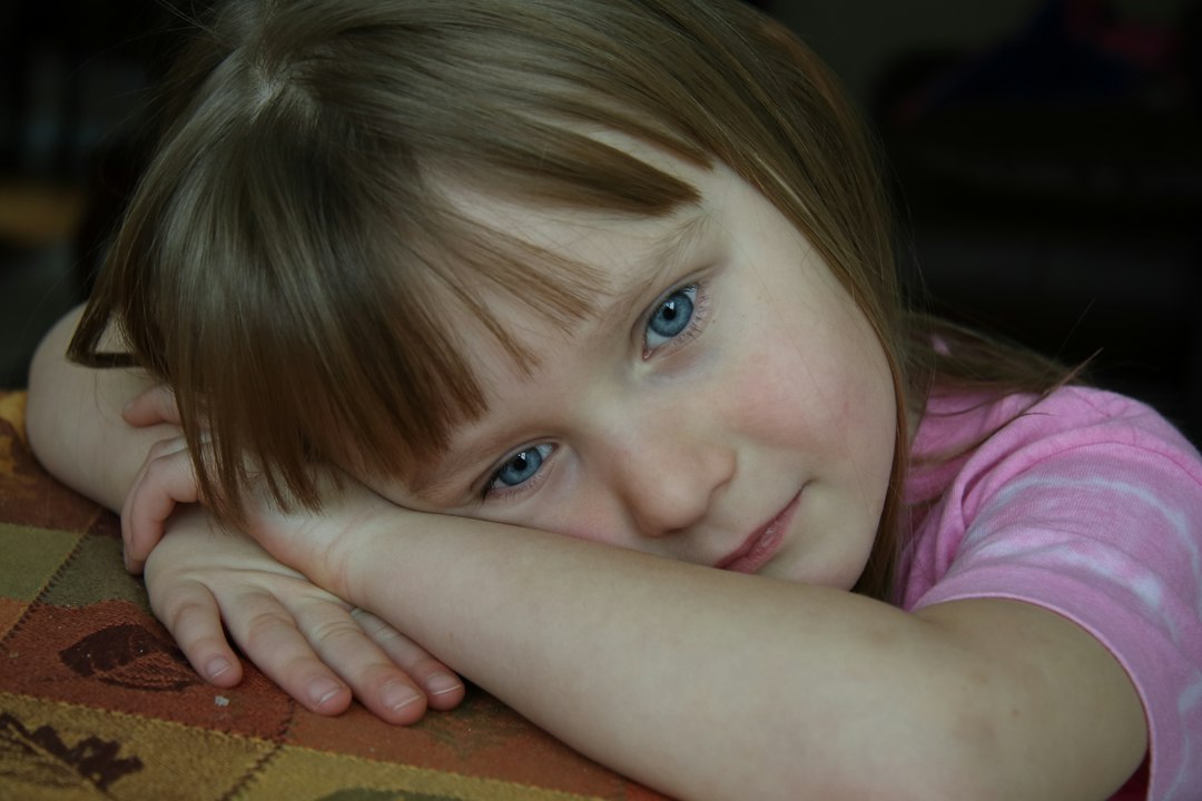 A girl sits at a table with her head resting gently on her folded hands, looking quiet and contemplative.
